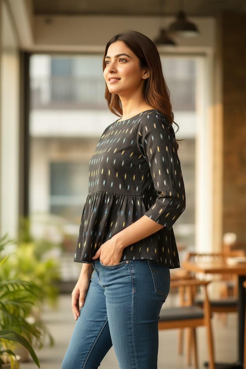 Naanu black ikat weave peplum top with boat neck and 3/4 sleeves, featuring white, yellow, and blue dash motifs. Model wears blue jeans.