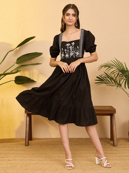 Naanu black cotton midi dress with white floral embroidery on bodice, square neckline, puff sleeves, and tiered ruffle hem.