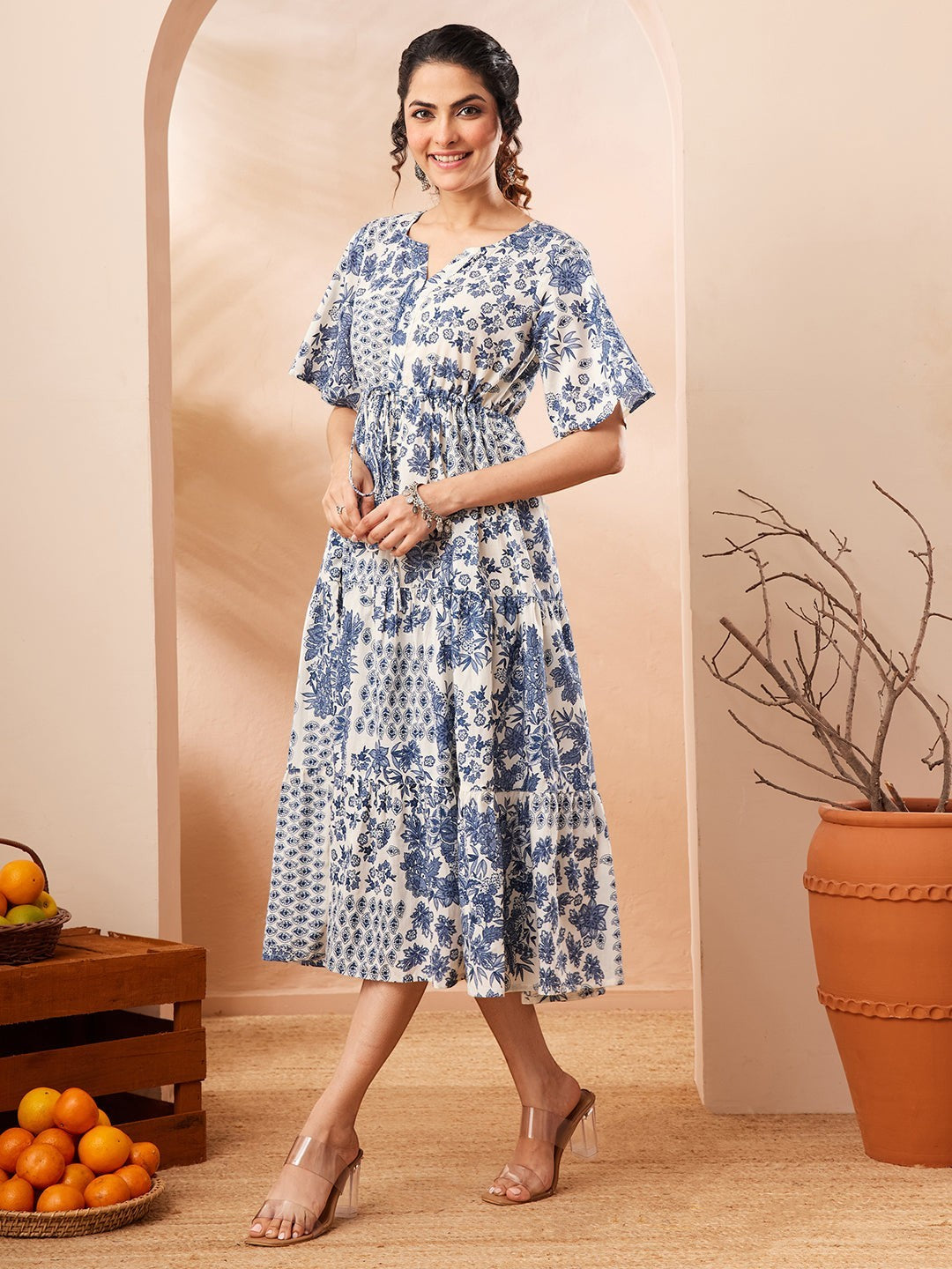 Naanu blue and white floral patchwork tiered midi dress with flutter sleeves and notched V-neck. Model wears clear heels.