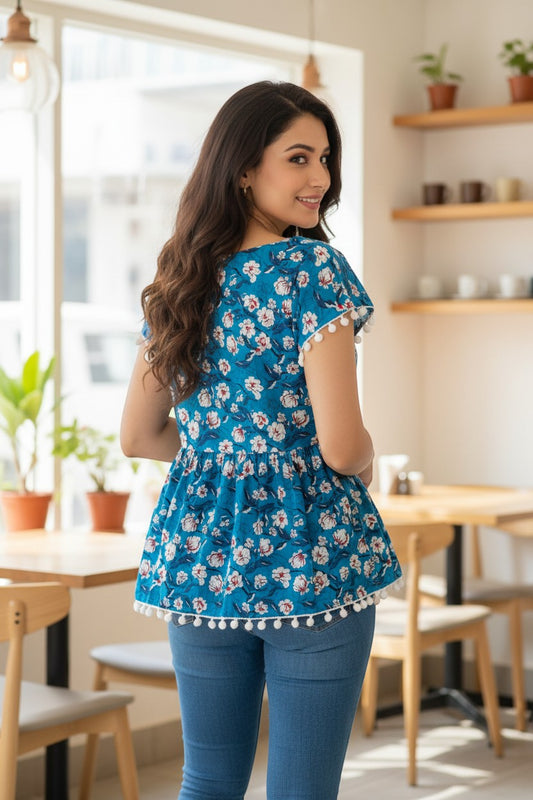 Naanu teal blue peplum top with white and pink magnolia floral print, cap sleeves, and white pom pom hem.