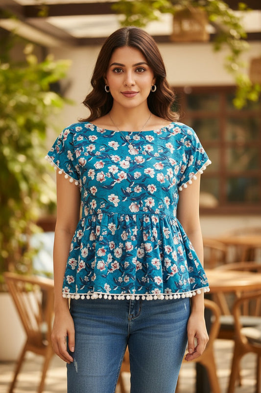 Naanu teal blue peplum top with white and pink magnolia floral print, cap sleeves, and white pom pom hem.
